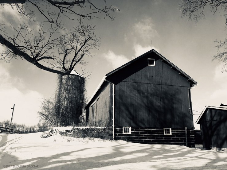 black and white photograph of a barn an silo on a snowy hill